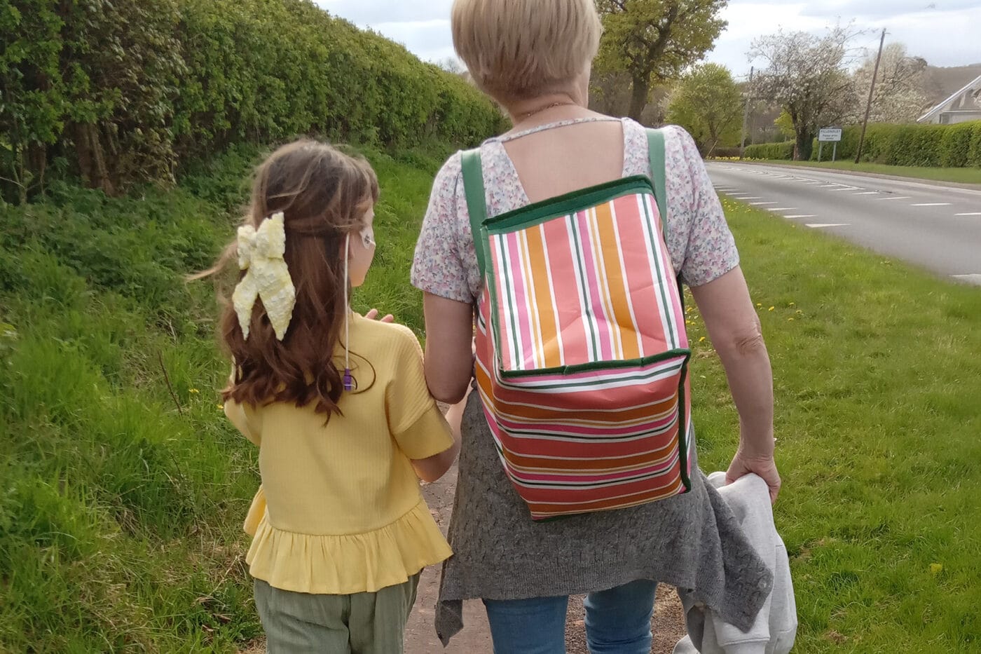 Child and adult walking along a roadside grassy verge, with a backpack and flower hair clip, representing support and guidance, young foundations, child welfare, family support, outdoor activity relevance.
